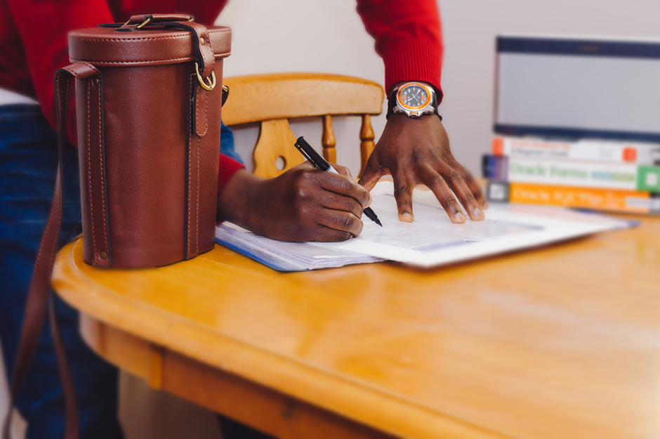 Man with watch on wrist signing contract on wooden table with leather bag and books