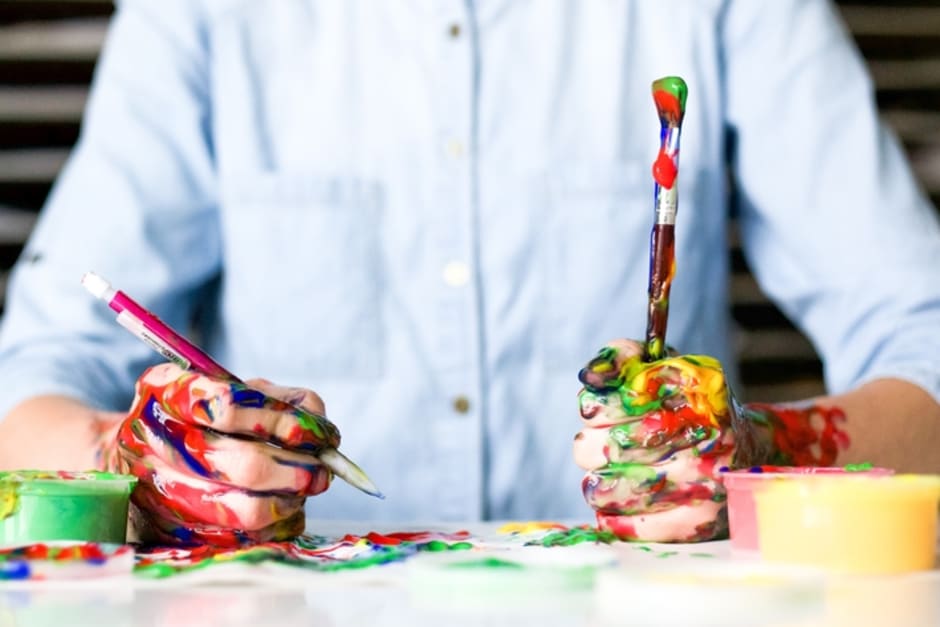 An image of an individual holding a pencil and paintbrush. The individuals forearm is covered in multicoloured paint splatters.