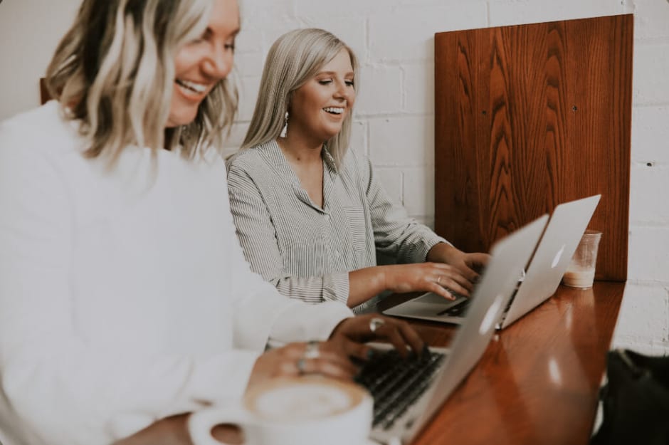zwei blonde Frauen arbeiten lachend an Laptops