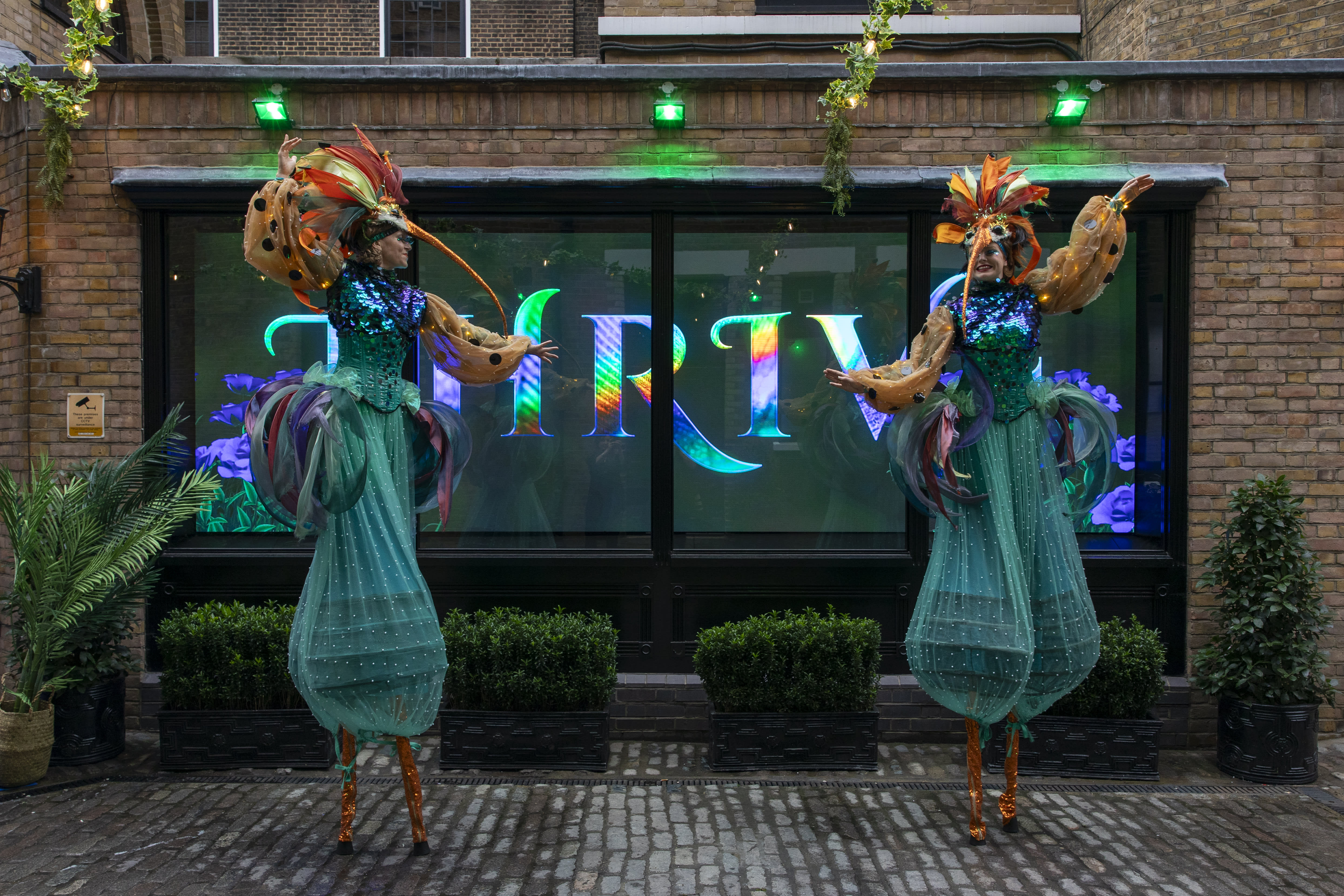 Two stilt-walking performers, dressed as hummingbirds, stand in front of a screen that reads 